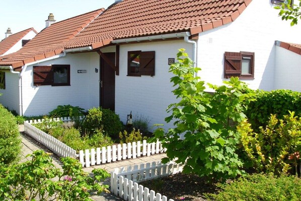 Un bâtiment blanc avec un toit recouvert de tuiles rouges, une clôture blanche en bois et un petit jardin agrémenté de différentes plantes.