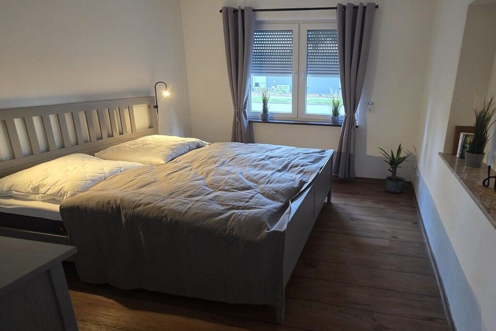 Ein Schlafzimmer mit einem Bett, einem Nachttisch, einem Fenster mit Vorhängen und einer Zimmerpflanze.