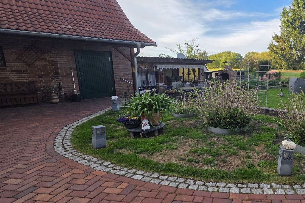 Ein Backsteinhaus mit einem Ziegeldach, einer überdachten Terrasse und einem angelegten Garten mit Blumentöpfen und einem kleinen Tisch.