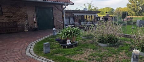 Ein Backsteinhaus mit einem Ziegeldach, einer überdachten Terrasse und einem angelegten Garten mit Blumentöpfen und einem kleinen Tisch.