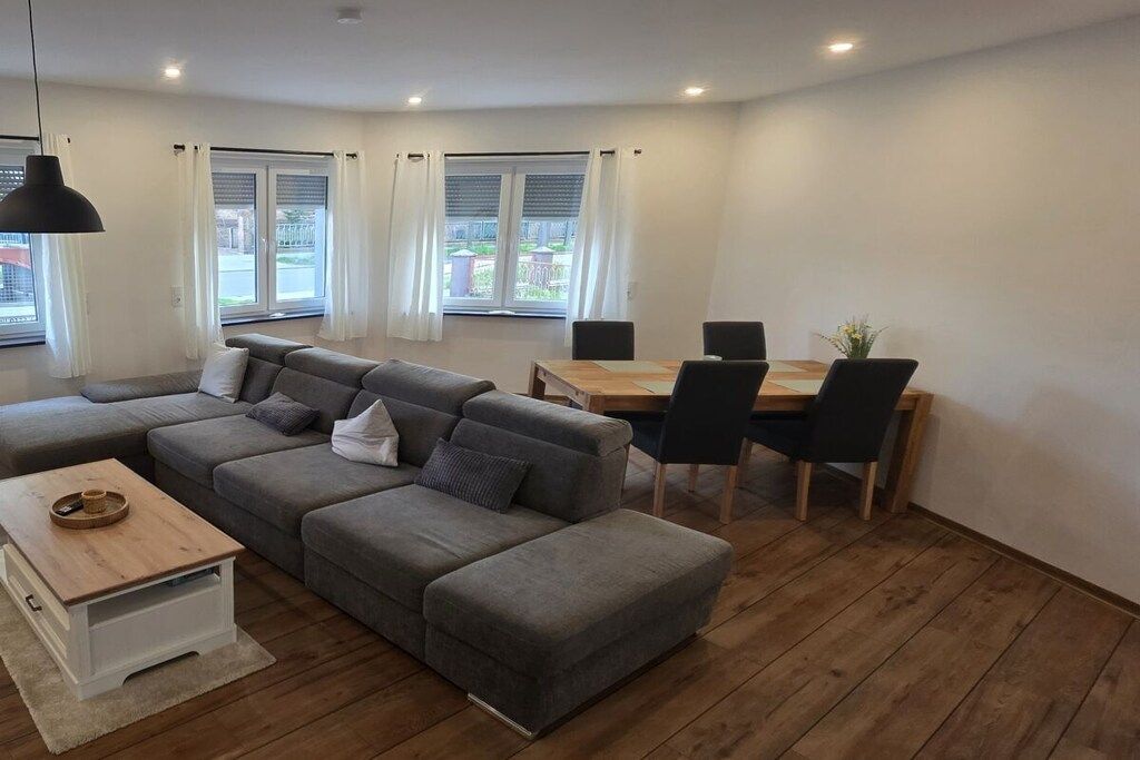 A modern living room with a grey sectional sofa, a wooden coffee table, and a dining area with a wooden table and chairs.