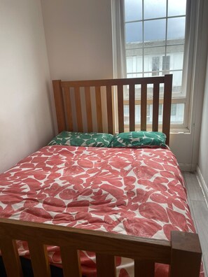 A small, single-bed room with a wooden headboard, a window with a view of buildings, and a patterned bedspread. - Charming 6-bedroom house in serene Erith perfect for families (Erith)