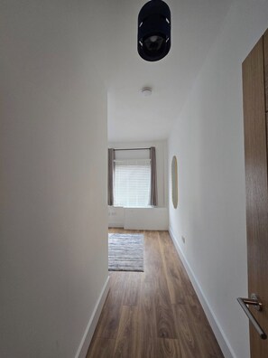 A hallway with wooden flooring, a door on the right, and a window with blinds in the distance. - Modern Apartment w/ Free Parking & Fast WiFi (Gloucestershire)