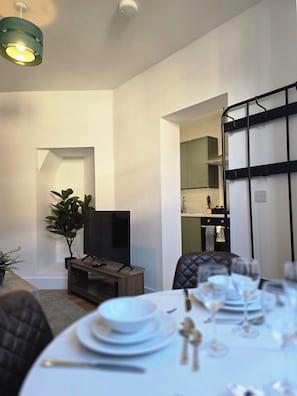 A modern living room with a flat-screen TV, a dining table set for four, and a kitchen area visible through an open doorway. - Modern Apartment w/ Free Parking & Fast WiFi (Gloucestershire)