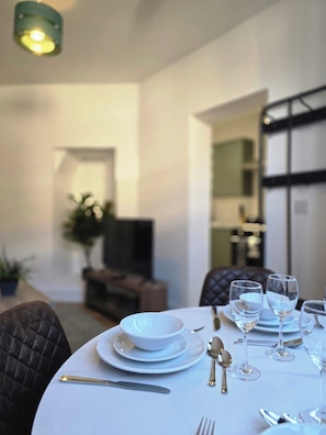 A dining table set with white plates, bowls, and cutlery, with a television and a plant in the background. - Modern Apartment w/ Free Parking & Fast WiFi (Gloucestershire)