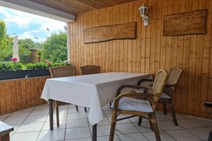 A covered patio with a table and wicker chairs, wooden walls, and a view of greenery. - Holiday home in Märkische Heide with balcony (Märkische Heide)