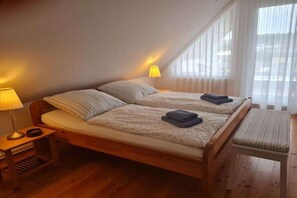 A bedroom with a wooden bed, two bedside lamps, a window with sheer curtains, and a small wooden stool. - Holiday home in Märkische Heide with balcony (Märkische Heide)