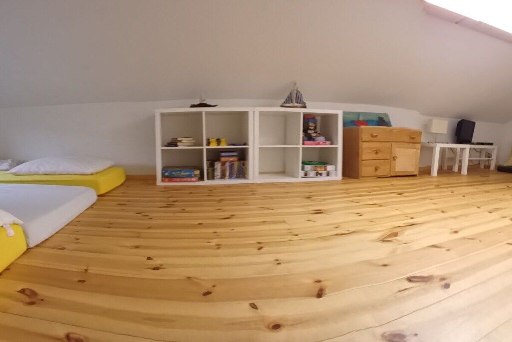 Ein Zimmer mit schrägem Dach, einem Bett mit gelbem Kopfteil, einem weißen Kleiderschrank, einem kleinen Tisch mit Lampe sowie einer weißen Regaleinheit mit Büchern und Kartons.