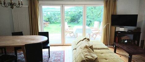 A living room with a sofa, a TV, a dining table with chairs, and a view through a glass door to an outdoor area.