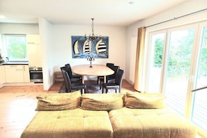 A modern living room with a sofa, a dining table with chairs, a chandelier, and a large window with a view. - Burkhardt Holiday Home (Neustrelitz)