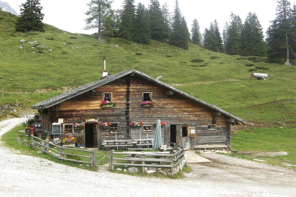 A rustic wooden chalet with a gravel driveway, surrounded by a wooden fence and a grassy hillside.