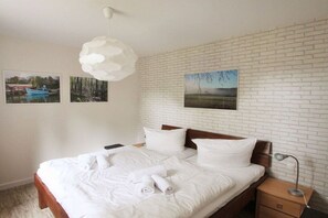 A hotel room with a white brick wall, a bed with white linens and towels, a wooden headboard, a nightstand with a lamp, and two framed pictures. - Holiday apartment with 1 bedroom (Ostseebad Sellin)