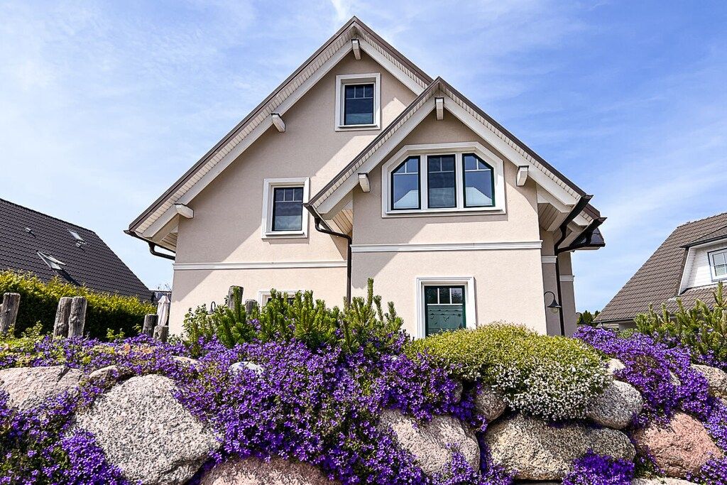 Ein zweistöckiges Haus mit Satteldach, einer Veranda und einem Garten mit violetten Blumen.