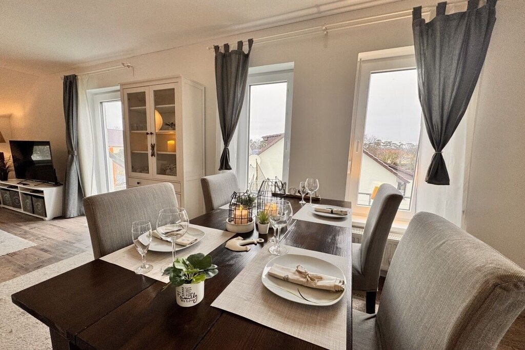 Un comedor con una mesa lista para cuatro personas, un televisor al fondo y vistas al exterior a través de amplios ventanales.