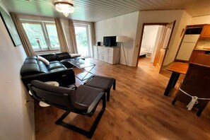 A modern living room with a sofa set, a coffee table, and a dining table. There is a television on a cabinet, and a lamp is visible. - Holiday apartment with 1 bedroom (Butjadingen)