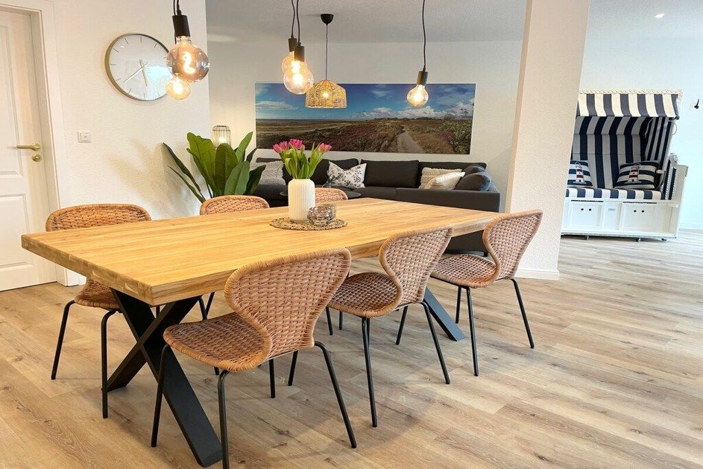 A dining area with a wooden table and wicker chairs, a clock on the wall, and a sofa with a striped cover in the background.