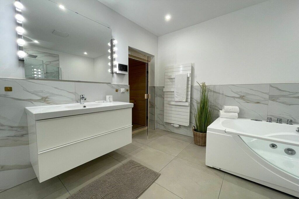 Ein modernes Badezimmer mit einem weißen Waschtisch, einem großen, beleuchteten Spiegel, einer Badewanne und einer Topfpflanze.