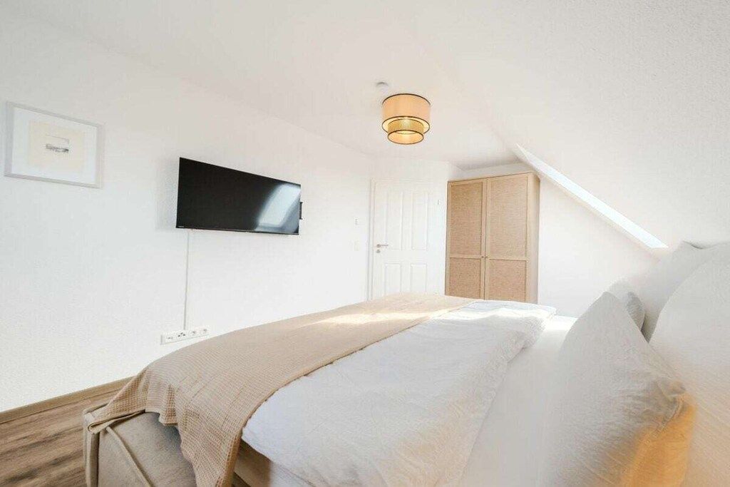 Quarto com cama, televisão na parede, luminária no teto e porta para outro cômodo.