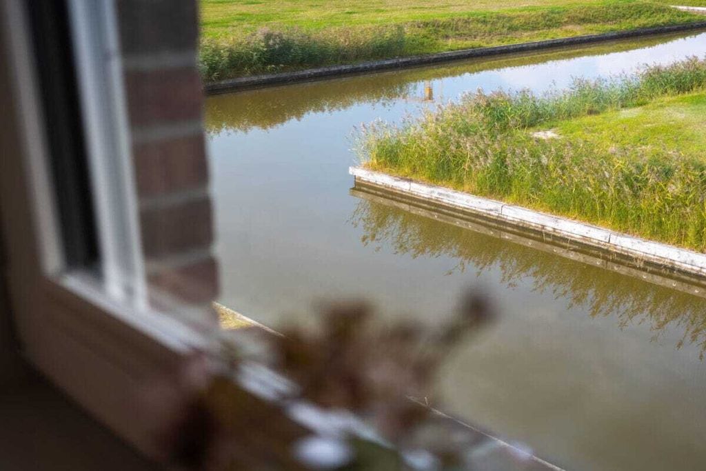 Une vue sur une étendue d’eau calme, bordée d’une rive herbeuse, que l’on aperçoit à travers une fenêtre donnant sur un mur de briques.