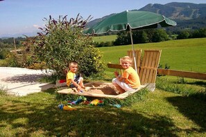 Zwei Kinder spielen im Sandkasten mit einem grünen Sonnenschirm und einem Holzstuhl in einer grünen, offenen Landschaft mit Bergblick.
