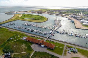An aerial view of a marina with boats docked, a large building with a red roof, and a curved dock extending into the water. - Holiday apartment with 1 bedroom (Norderney)