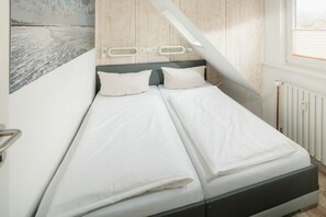Ein Bett mit weißer Bettwäsche, ein Kopfteil mit heller Holzoptik und ein Fenster mit Jalousien.