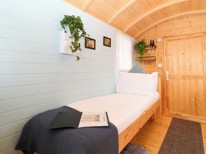 Un lit simple avec une couverture sombre et un guide d’accueil, une porte en bois et une petite étagère avec des bouteilles.