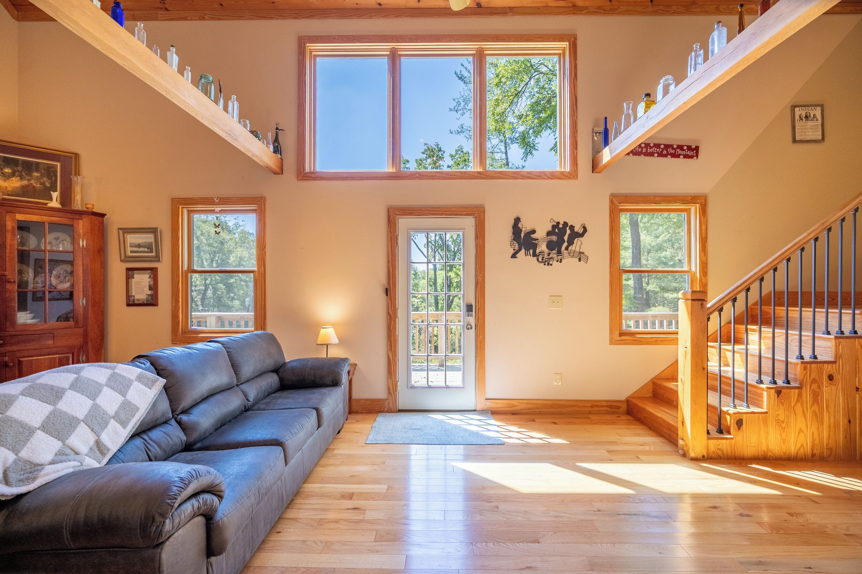 A spacious living room with a leather sofa, a wooden staircase, and multiple windows offering views of the outdoors.