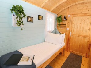 Ein Zimmer mit einem Einzelbett, Holzboden, einem Fenster mit Vorhängen, einem Nachttisch mit einem Buch und einer Tür mit einer Pflanze daneben.