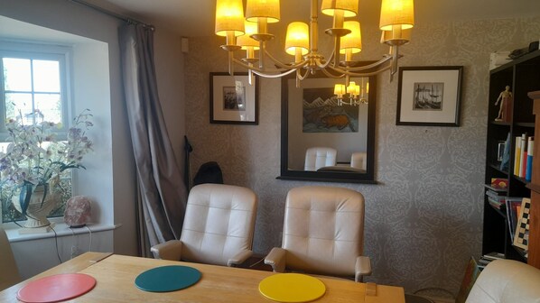 A dining area with two chairs, a table with colorful placemats, a chandelier, framed artwork on the wall, and a bookshelf with books and decorative items.