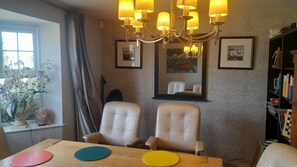 A dining area with two chairs, a table with colorful placemats, a chandelier, framed artwork on the wall, and a bookshelf with books and decorative items.