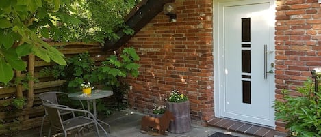 Ein Ziegelhaus mit einer weißen Tür, einer kleinen Terrasse mit Tisch und Stühlen und Blumentöpfen.