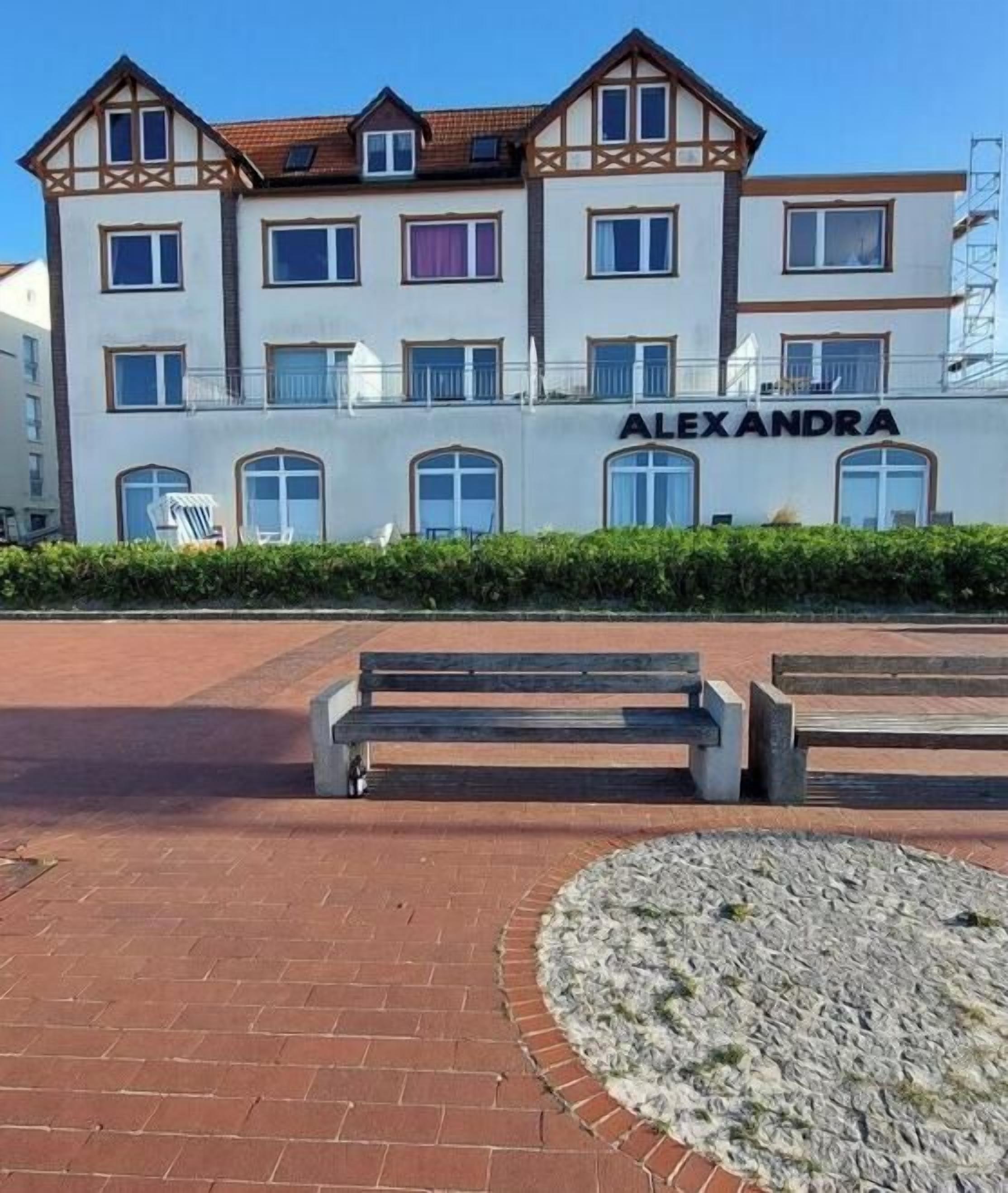 Un edificio de varios pisos con el nombre 'ALEX ANDRA' en la fachada, que presenta un diseño simétrico con un balcón central y un suelo de ladrillo.