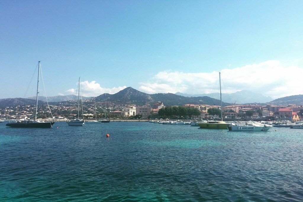A coastal marina with boats docked, a clear sky, and a mountainous backdrop.