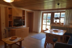 Ein gemütliches Wohnzimmer mit Fernseher, Holzschränken, einem Esstisch mit Stühlen und einem großen Fenster mit Vorhängen.