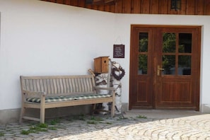 Un banc de fusta amb coixí de vares a l'exterior d'un edifici amb porta de fusta i un rètol fixat a la paret.