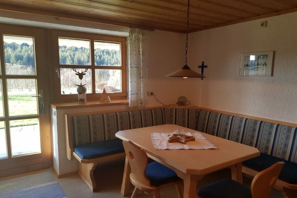 A room with a wooden table and benches, a window with a deer head decoration, and a cross on the wall.
