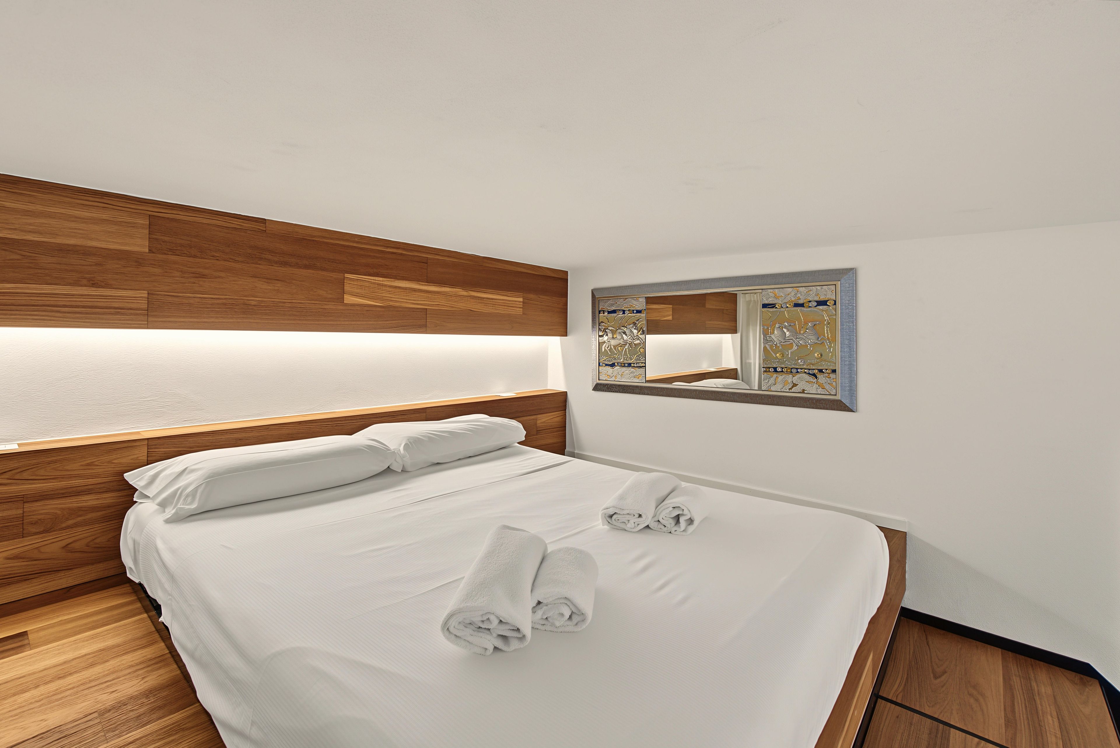 A neatly made bed with white linens and towels, a wooden headboard, and a wall-mounted mirror with a decorative frame.