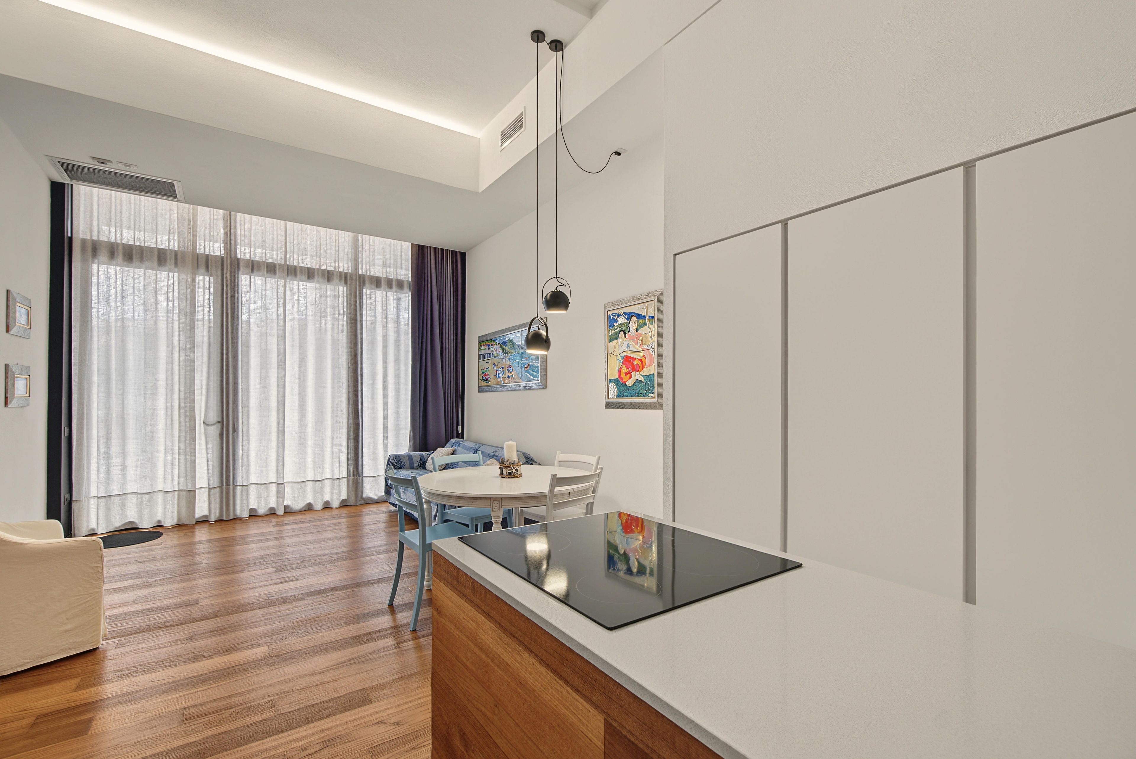 A modern kitchen with a central island, a dining area with a round table, and a living space with a sofa and curtains.