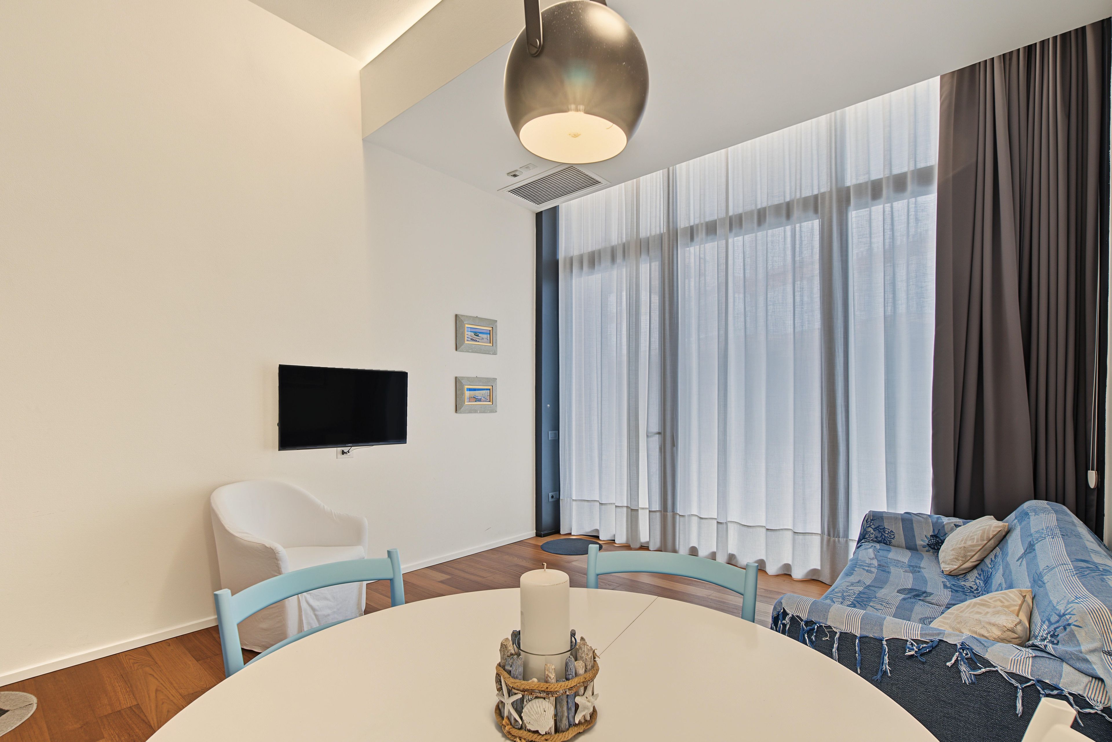 A modern living room with a round table, a flat-screen TV mounted on the wall, a sofa with patterned cushions, and a large window with sheer curtains.