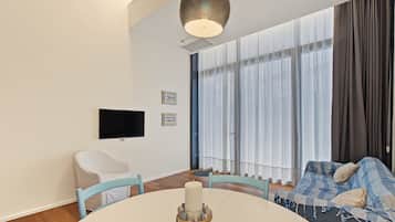 A modern living room with a round table, a flat-screen TV mounted on the wall, a sofa with patterned cushions, and a large window with sheer curtains.
