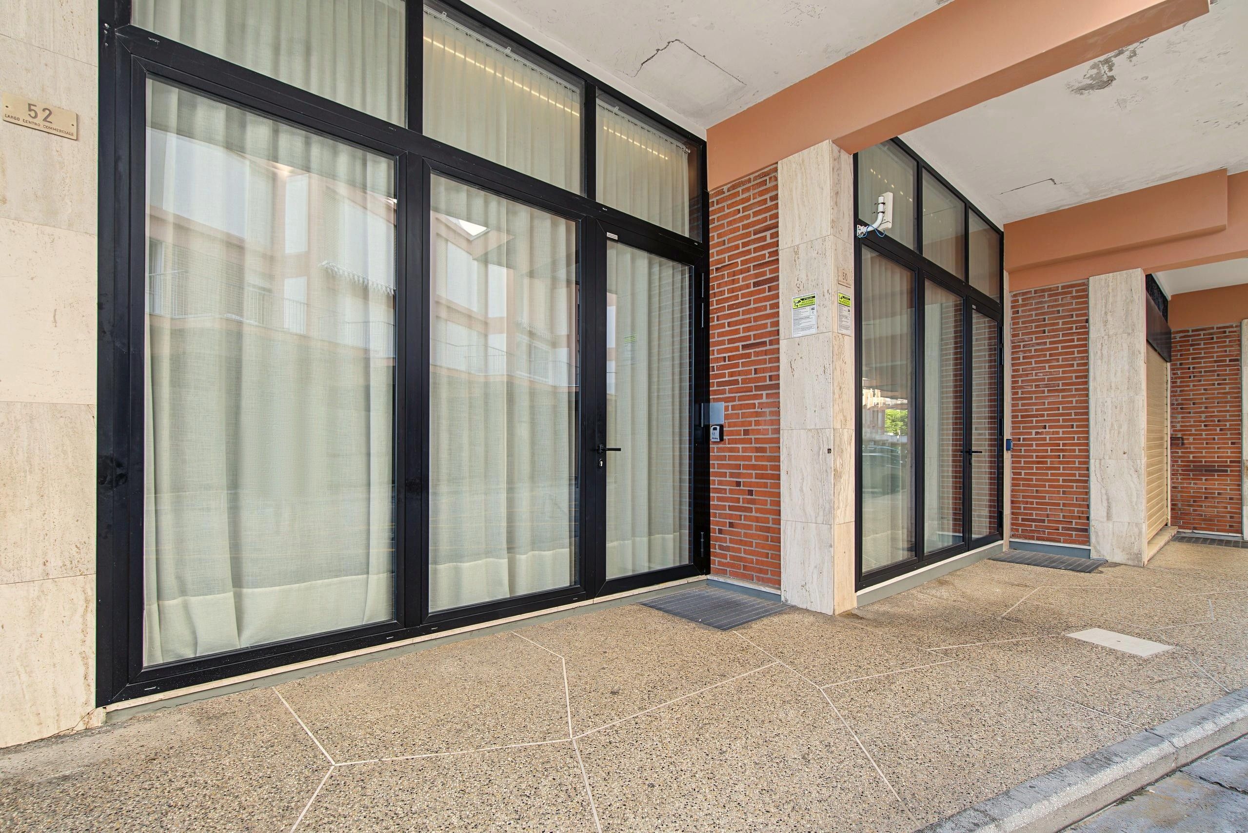 Fachada de un edificio con amplias puertas y ventanas de vidrio, un muro de ladrillo y suelo de baldosas.