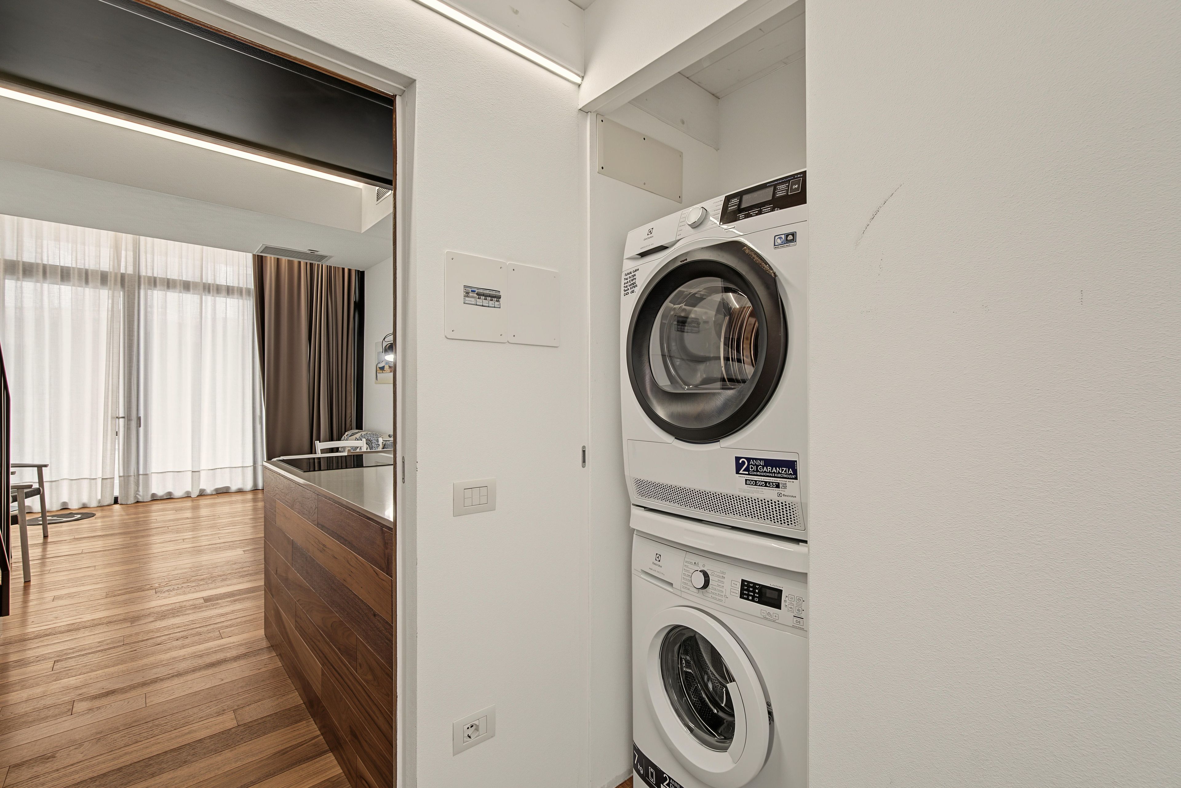 Un lavadero con lavadora y secadora de carga frontal, suelo de madera y vista a una sala de estar con sofá y cortinas.