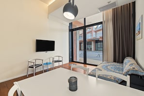 Una camera d'albergo moderna con una grande finestra, una TV a schermo piatto, un tavolo da pranzo e un divano con cuscini decorati.