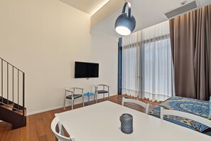 Una moderna camera d'albergo con TV a schermo piatto fissata al muro, un tavolo da pranzo con sedie, un piccolo tavolino blu e un letto con biancheria a motivi decorativi.
