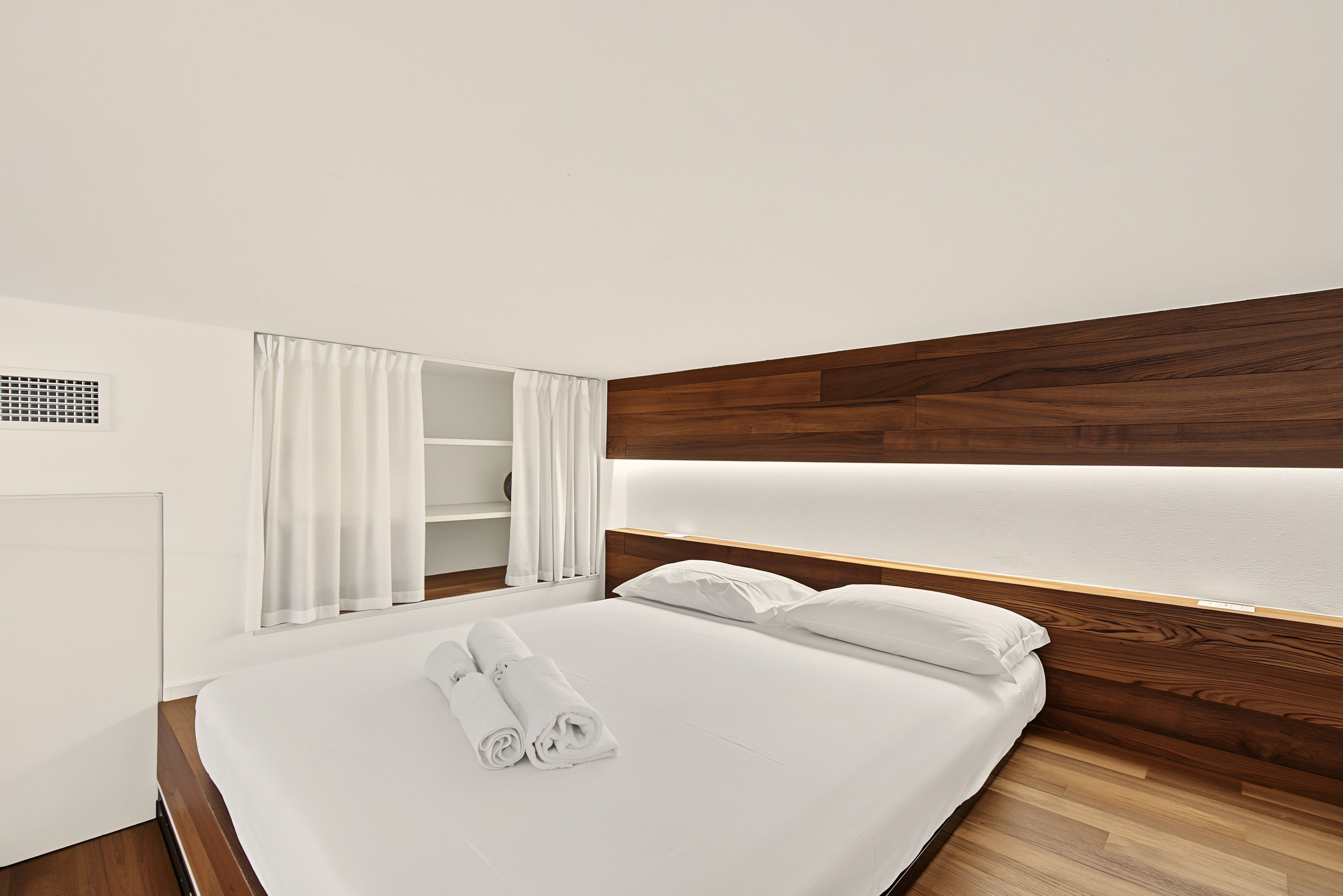 Una camera d'albergo con un letto rifatto, una testiera in legno, un comodino bianco e un armadio bianco con uno specchio.