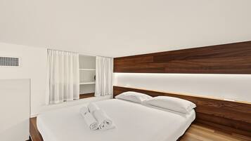 Una camera d'albergo con un letto rifatto, una testiera in legno, un comodino bianco e un armadio bianco con uno specchio.