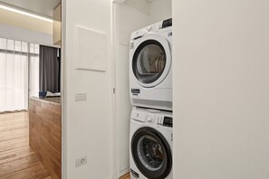 Un lavadero con dos lavadoras apiladas, piso de madera y una puerta que da a otra habitación.