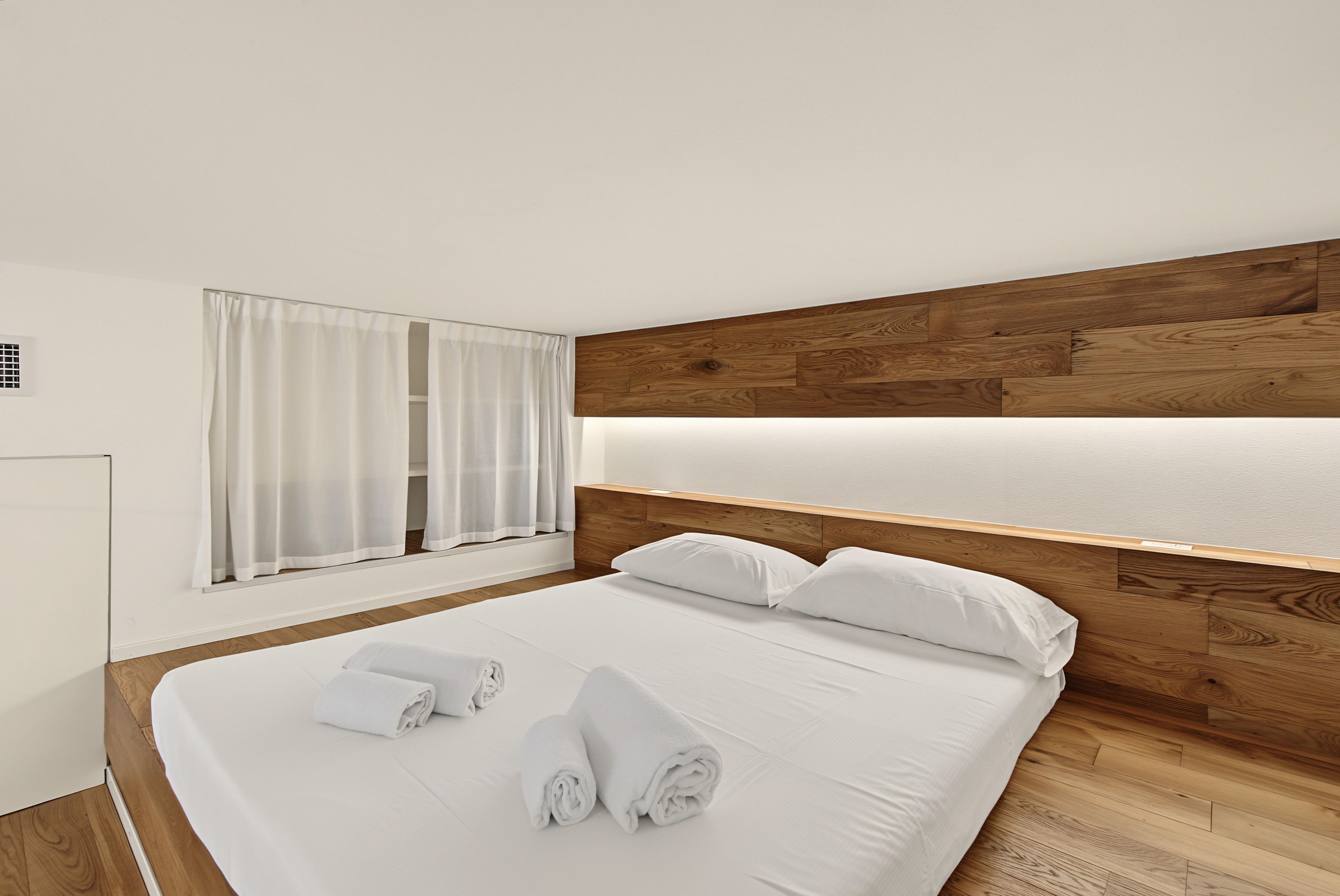 Una camera d'albergo con una testiera in legno, un letto grande con biancheria da letto bianca e una finestra con tende bianche.