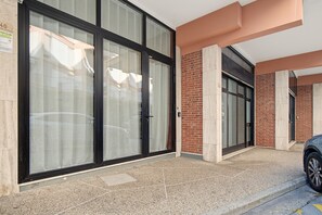 Fachada de edificio con grandes puertas de vidrio, pared de ladrillo y zona de estacionamiento con un coche.